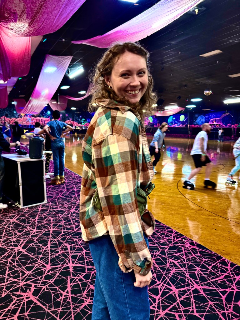A young woman with curly brown hair stands in profile. Her head is turned towards the camera with a smile on her face. In the background is a vibrant roller rink.